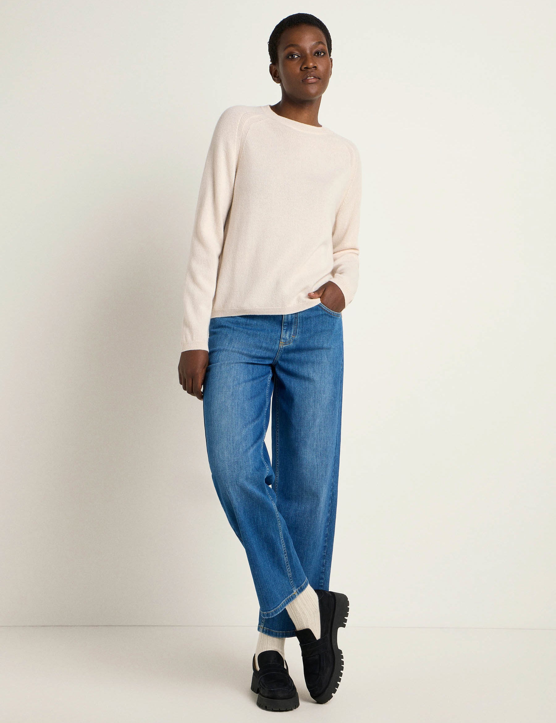 Frau in beigem Pullover und blauen Jeans steht