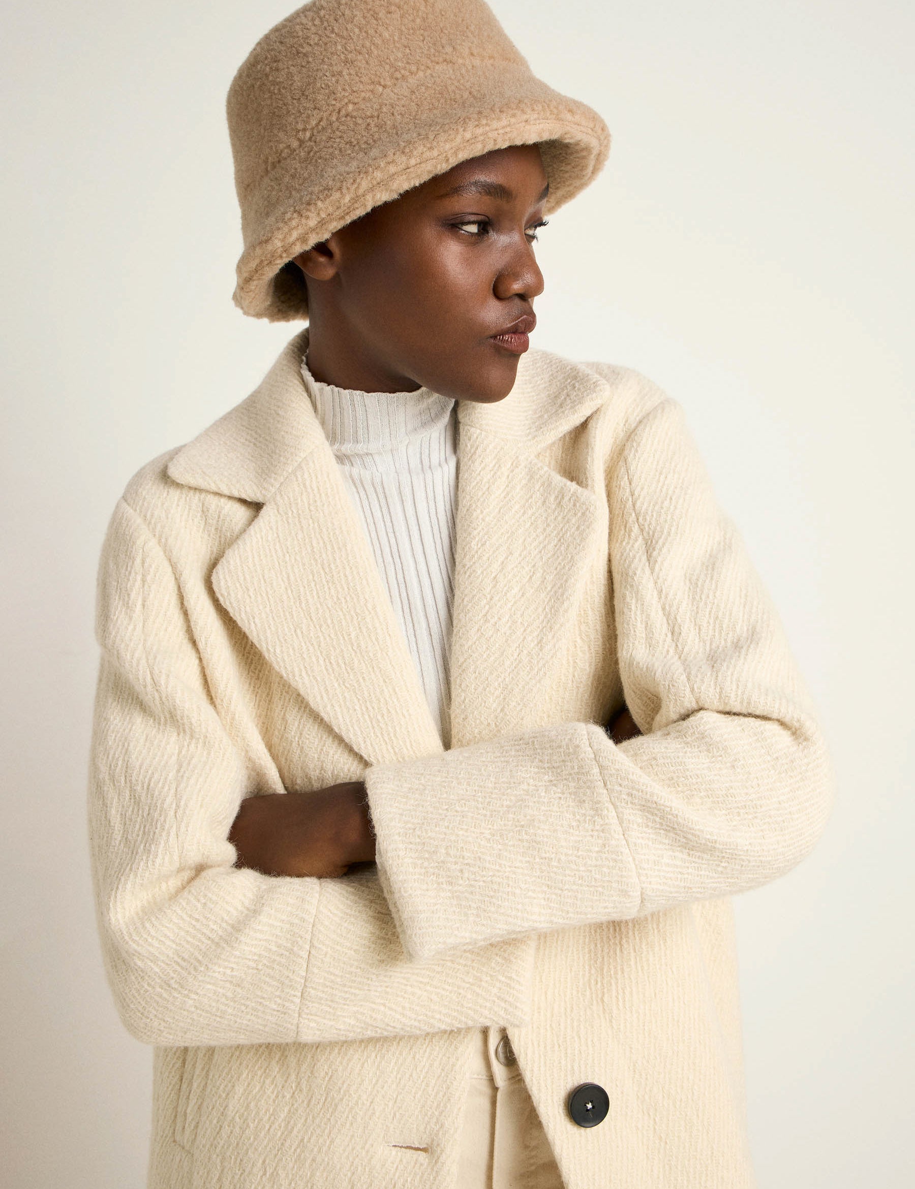 Frau in cremefarbenem Mantel und Hut schaut nachdenklich