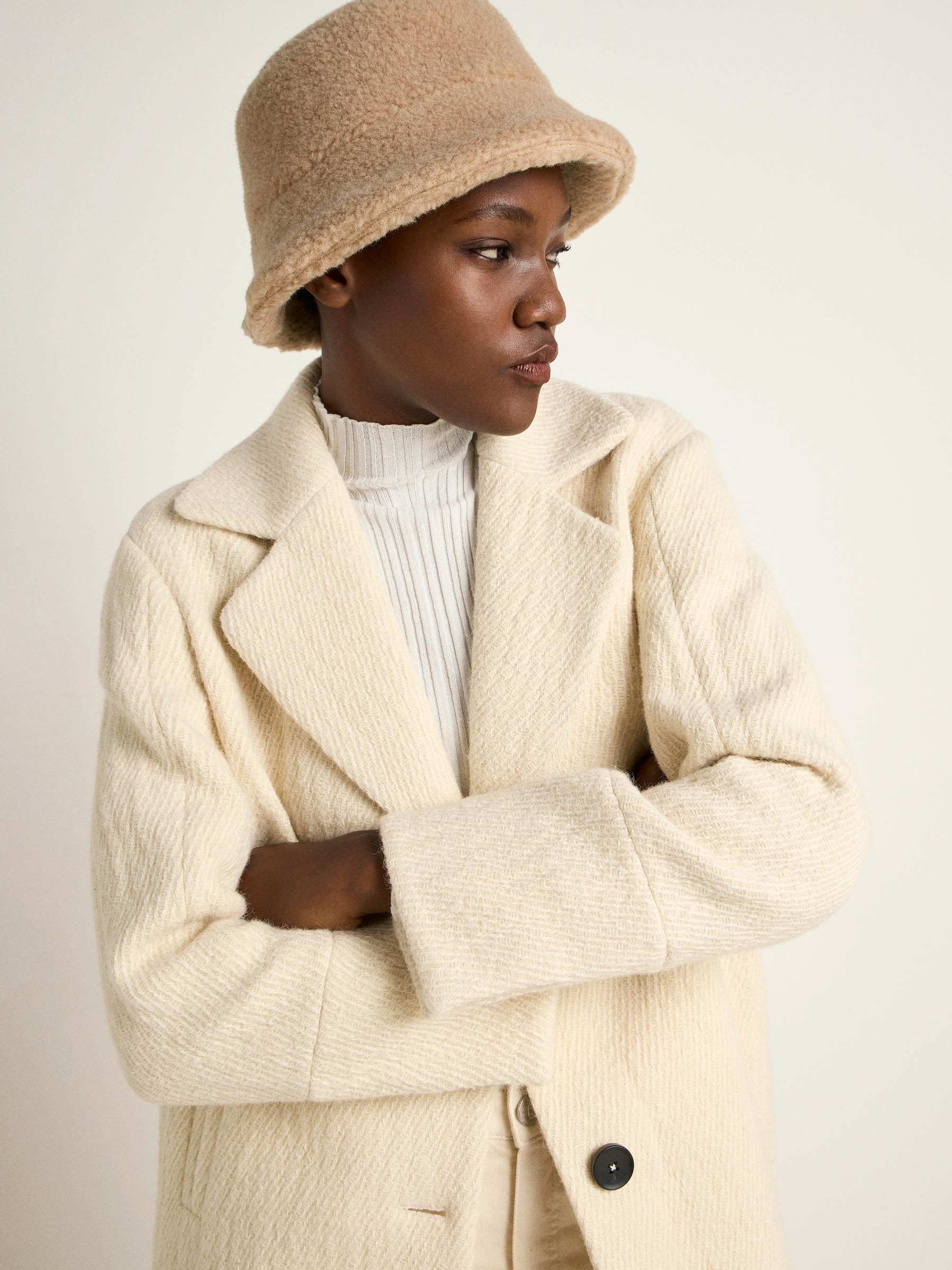 Frau in cremefarbenem Mantel und Hut schaut nachdenklich