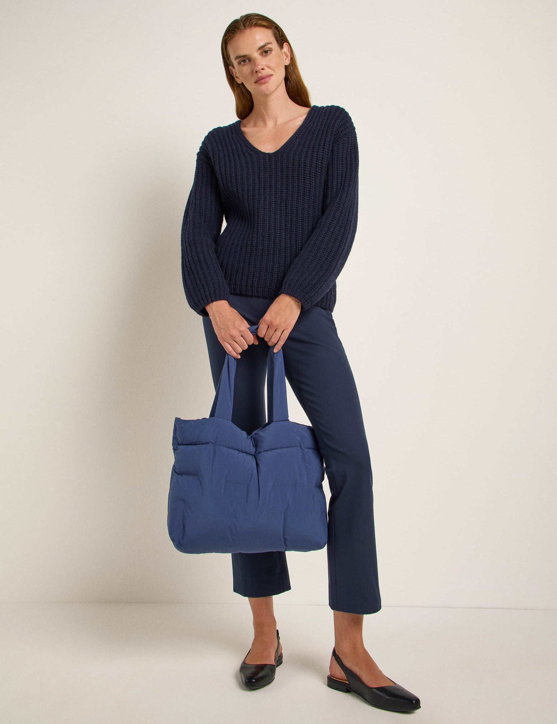 Frau mit Pullover, Hose und großer Tasche posiert.